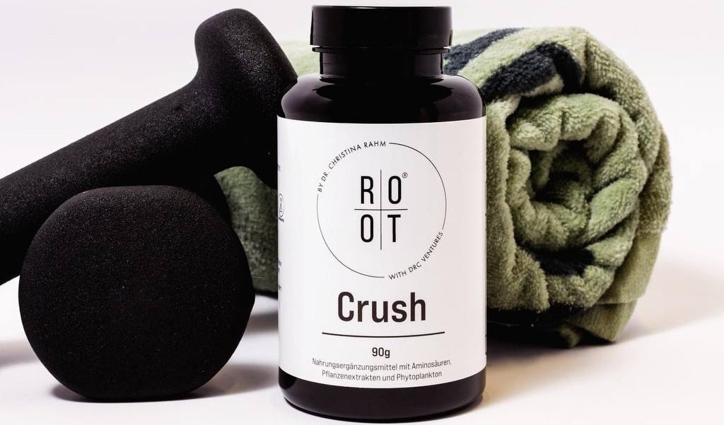 Root crush: Hanteln, ein grünes Handtuch und in der Mitte klar und deutlich eine Dose Root Crush mit 90gr. Inhalt